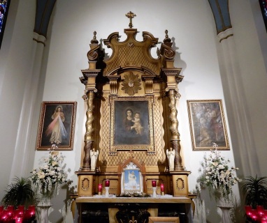 close-up view of the Madonna & Child over the altar in the left alcove: lit candles on either side at the forefront of the altar, floral arrangements on either side of the altar, & framed pictures on the wall (left: Divine Mercy; right: baptism of Jesus)