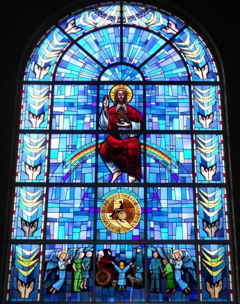 Stained-glass window of the Sacred Heart of Jesus seated on a rainbow with the words “YOUR HANDS” on the left and “ARE MY HANDS” on the right