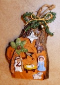 Painted wood ornament of the Nativity setting with the Holy Family, a donkey, the Star, a palm tree, & greenery with a tiny gold-colored bell and cord at the top.