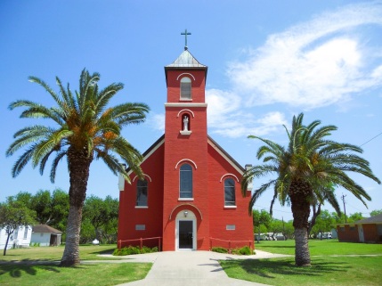 Rural setting church with a tall palm tree on either side of the entrance. Below the steeple, which runs from the cross at the top to the doorway on the ground level, are two windows above and below a four- or five-foot statue of Our Lady of Consolation. Above the front doors is a tallish window with similar ones on either side for a total of three, with the ones on the right and the left positioned about a foot lower on either side.