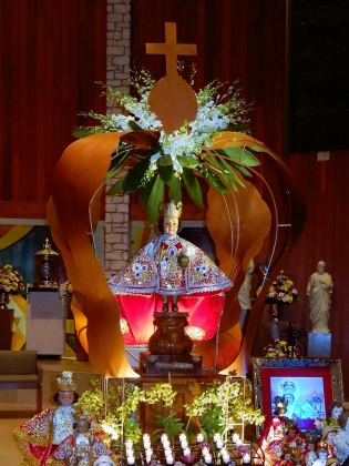 Church setting with an ornate statue of Santo Nino de Cebu enclosed in golden-brown hollowed-out crown enclosure with a with a cross on top & lit candles & smaller statues on the table below in the foreground