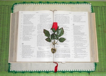 Faux rose--- leaves on stem from which is attached a small heart with a cross on its center--- rests on the center of an open Bible which rests atop a linen cloth with an embroidered edge all around on a wicker background