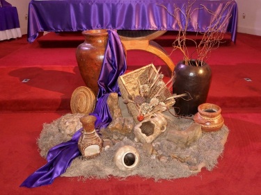 On the floor before the Lenten altar is an earth-colored cloth topped with fabric like hay. On that are various gourds and jugs with twigs emanating from the large one on the right and, from the taller one on the left, a long satin cloth like water flowing from a waterfall through the ceramic containers below it.