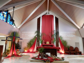 Palm Sunday altar with empty jugs on a mat on the floor in front of the altar and a ceiling-to-floor curtain with narrow purple curtain on either side on the back wall. To the left are three full-sized plain, dark-wood crosses--- the tallest in the center draped in red--- with palm fronds and ceramic containers around them on the floor. Palm fronds adorn the back wall, too.